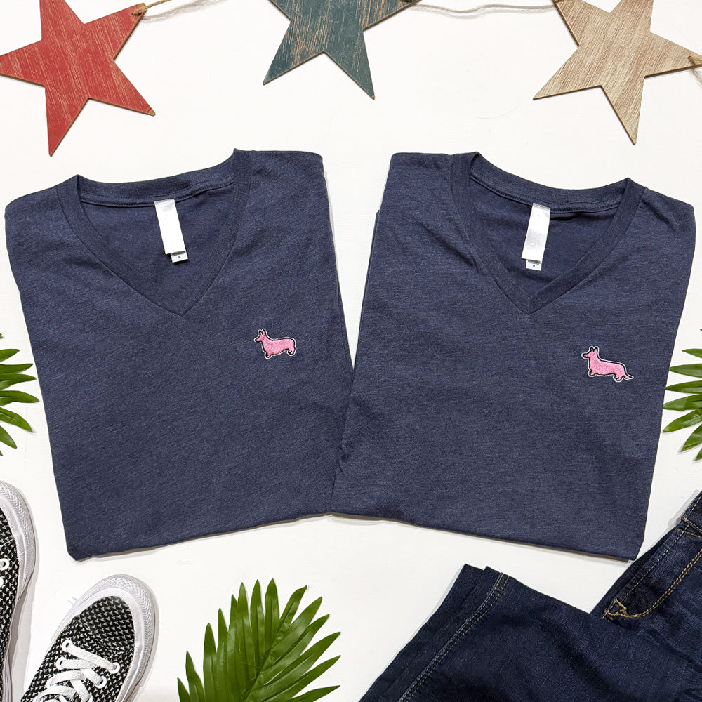 Two folded navy blue V-neck shirts with pink dog logo on a white background with decorative stars and leaves.