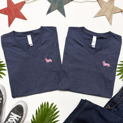 Two folded navy blue V-neck shirts with pink dog logo on a white background with decorative stars and leaves.